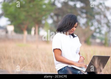 young beautiful black woman sitting alone outside in a park using her laptop Stock Photo