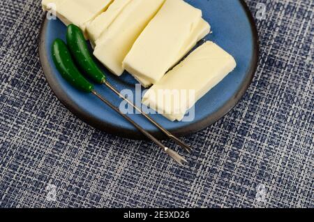Rectangular slice of processed cheese in a blue saucer on a denim background. Sliced into rectangular portions. Skewer for stabbing. Dairy products, f Stock Photo