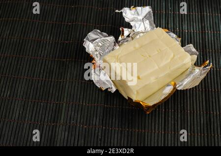 Rectangular piece of processed cheese in open foil package on a dark bamboo background. Cheese ready to eat. Dairy products, food. Stock Photo