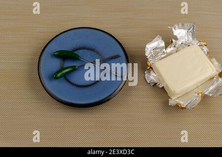 Rectangular piece of processed cheese in an open foil package on a brown background. Skewers for stabbing in a blue saucer. Cheese ready to eat. Dairy Stock Photo