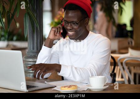 Afro hipster man wear red hat talking on cellphone remote online working on laptop computer in cafe Stock Photo