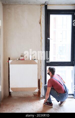 Plumber installing water radiator in new empty room next to window. Fitting lower valve. Male worker during installation process. Construction, maintenance, home refurbishment concept Stock Photo