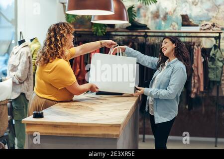 Friendly seller of clothing store giving shopping bags to satisfied female customer. Fashion store owner handing over the shopping bag to a female cus Stock Photo