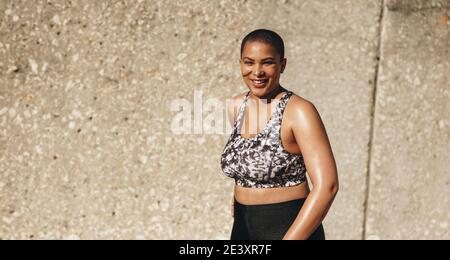 Portrait of a happy woman in sportswear standing by a wall outdoors and smiling. Woman with buzzcut hairstyle relaxing after workout. Stock Photo