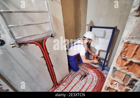 Wide angle photography of young plumber working in unfinished apartment, installing flush tank. Man wearing blue uniform and white helmet. Red pipes on the floor, new hanging radiator on the wall Stock Photo