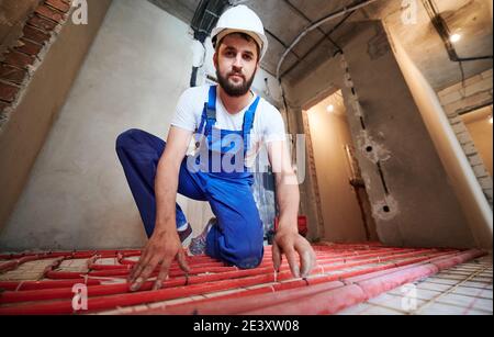 Wide and low angle view portrait of young plumber wearing blue overalls and white helmet, tying up red tubes on the floor heating system in new unfinished apartment. Concept of home renovation. Stock Photo