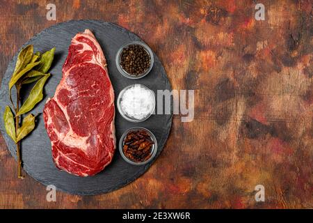 Raw beef ribeye on a round slate plate with glass containers containing peppercorns, coarse salt and cayenne chillies on an old wooden table in brown Stock Photo