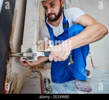 Bearded young man in work overalls using plumber wrench while installing heating radiator in room. Male plumber in safety helmet installing heating system in apartment. Focus on hand with instrument. Stock Photo