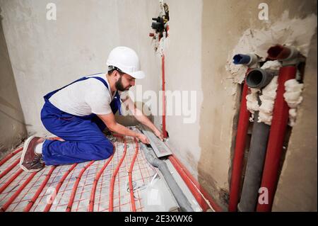 Wide angle view on young worker standing on his knees in blue overalls and helmet installing underfloor heating system and plumbing system. Concept of home renovation and plumbing works. Stock Photo