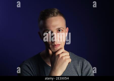 Young caucasian blonde man in grey t shirt on dark purple background touching his chin and thinking about something. Stock Photo