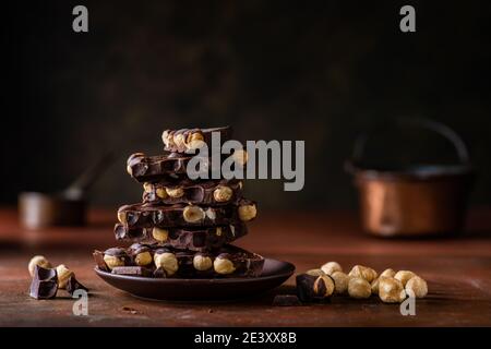 In the foreground some stacked pieces of dark chocolate with hazelnuts. Composition and dark brown background. Still life Stock Photo
