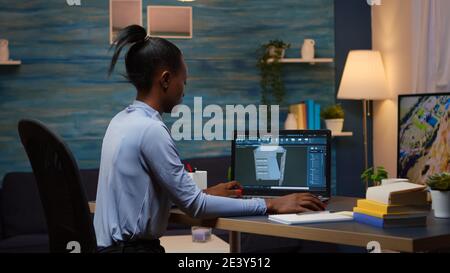 African lady enginer analysing digital mechanism of container checking prototype of project looking on laptop sitting at desk in living room office late night. Black employee using modern technology Stock Photo