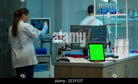 Microbiologist working on monitor in background analysing test samples with tablet with green chroma key screen placed on desk in front. Team of biotechnology scientists developing drugs. Stock Photo