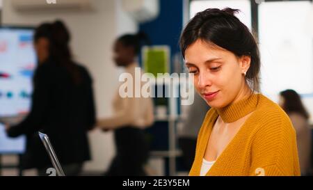 Young woman in casual clothing working at laptop sitting at desk in busy financial startup company while diverse team analyzes statistics data in modern office. Multiethnic team concentrated on work Stock Photo