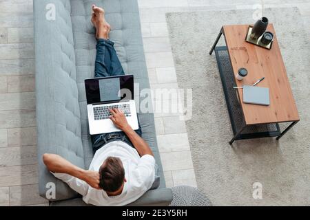 male freelancer is working on a laptop while lying on the couch . Stock Photo