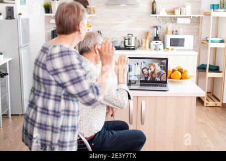 Over the sholder shot of grandparents waving on laptop, talking with family sitting in kitchen, communicating with niece via videocall. Parents and grownup daughter talking using videoconference app concept Stock Photo