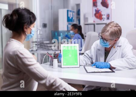 Elderly doctor holding tablet with green screen in clinic room speaking with young patient explaining the treatment looking at mockup display. Showing at izolated monitor, chroma key desktop. Stock Photo