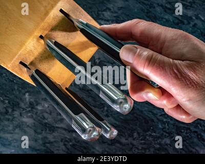 Closeup POV overhead shot of a man’s hand removing or replacing a cutting and chopping steel kitchen knife from its wooden holding block. Stock Photo