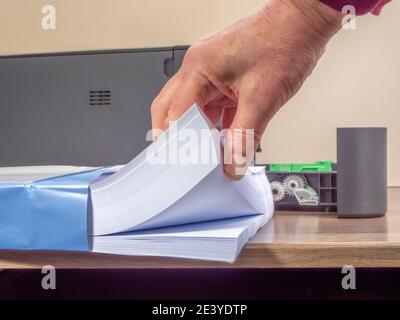 Closeup of a man’s hand holding the end of a ream of white paper, pulled halfway out of a new pack, in front of an open printer tray on a desk. Stock Photo