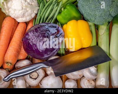 Closeup POV overhead shot of a steel cleaver lying among a variety of fresh, raw vegetables, waiting to be cut / chopped. Stock Photo