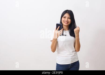 Overjoyed asian woman celebrating success with smartphone isolated on white, received good news, clenching fist with excitement, copy space Stock Photo