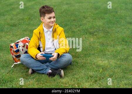 Schoolboy sits on the lawn with  smart phone in his hands. Modern teenager in yellow jacket Stock Photo