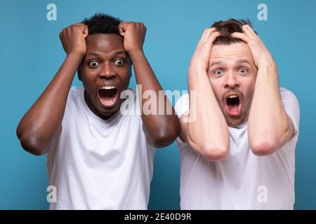 African and caucasian men feel scared afraid, confused and shocked by unexpected news. Studio shot on blue wall Stock Photo