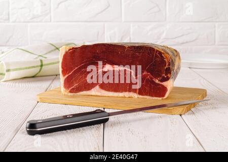 Jamon 1 kg piece and sharp long blade knife on a cutting board over white wooden table. Tasty spanish jamon Serrano. Traditional cured meat delicacy. Stock Photo