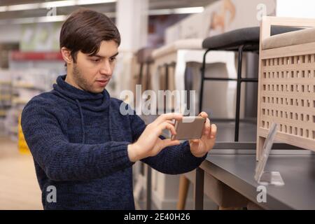 Handsome man using smart phone at home furnishings store, taking photo of a price label. Male customer choosing furniture for his new apartment, using Stock Photo