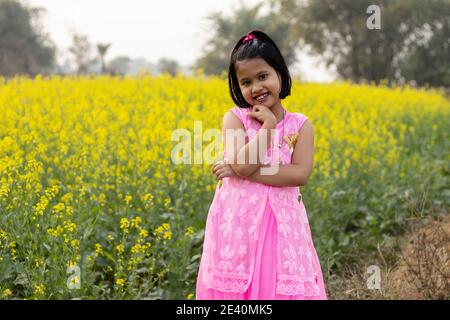 a pretty indian girl child in pink dress posing with smiling face near yellow mustard flower field Stock Photo