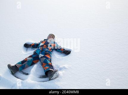 Joyful boy in winter overalls lying on the snow, moving his hand up and down, playing outdoors, enjoying the winter. A child in the snow shows the fig Stock Photo