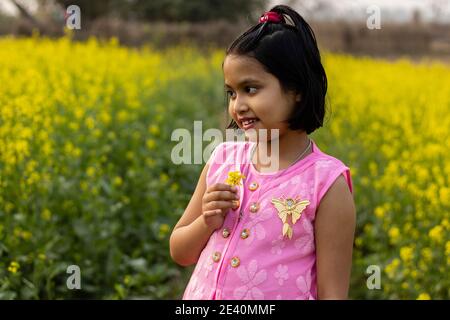 a pretty indian girl child in pink dress standing with a mustard flower in hand near yellow mustard flower field Stock Photo