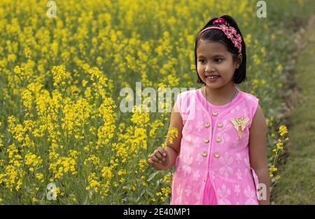 a pretty indian girl child in pink dress standing near yellow mustard flower field Stock Photo