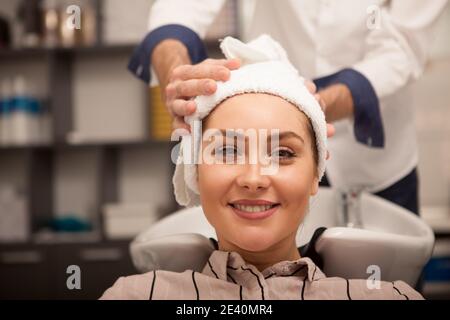 Close up of a happy beautiful woman with her hair wrapped in a towel after washing at beauty salon. Charming female smiling joyfully waiting for a new Stock Photo