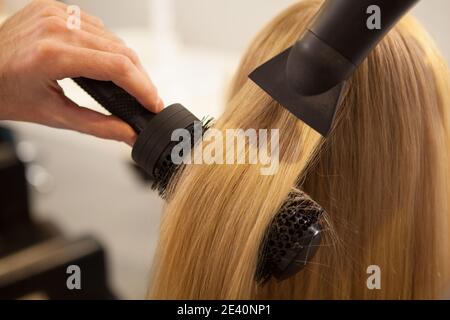 Cropped close up of a professional hairdresser using round brush and blow dryer, styling long blond hair of his female client. Healthy strong hair of Stock Photo