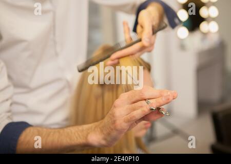 Cropped close up of a professional hairdresser working at his salon, cutting hair of a blond female client. Hairstylist using comb and scissors, worki Stock Photo