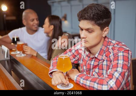 Young depressed man feeling lonely, drinking alone at the bar, affectionate couple cuddling on background Stock Photo