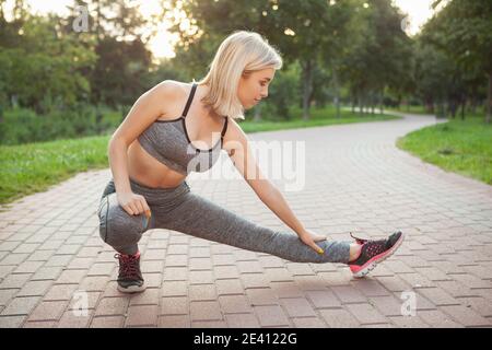 Blond haired young sporty woman stretching at the park before running. Attractive athletic woman exercising outdoors in the morning, copy space. Healt Stock Photo
