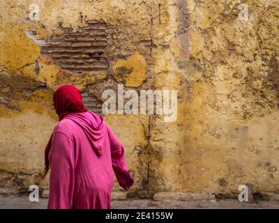 A woman dressed in typical Arab clothing with strong colors, walking in front of an old facade of a square in Morocco. Stock Photo