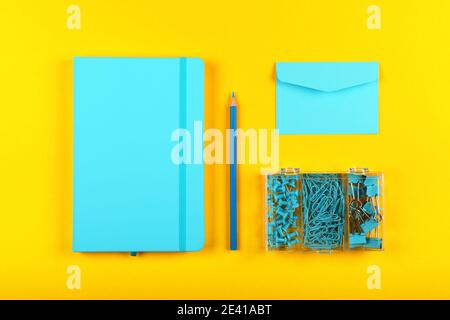 Neatly organized stationery flat lay of pastel blue notebook, envelope, pencil, office clips, binders and pins in box over yellow Stock Photo