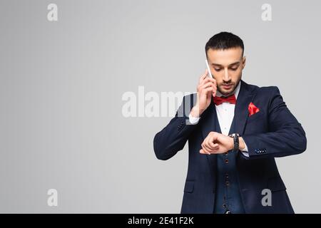 elegant hispanic man checking time on wristwatch while talking on smartphone isolated on grey Stock Photo