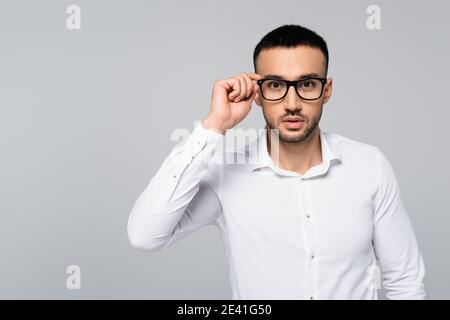 young hispanic manager in white shirt adjusting eyeglasses while looking at camera isolated on grey Stock Photo