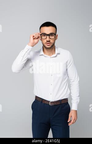 brunette hispanic businessman adjusting eyeglasses while looking at camera isolated on grey Stock Photo