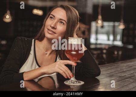 Lovely attractive woman relaxing at the pub, having delicious beer, looking away dreamily. Charming beautiful woman resting, drinking beer at the bar, Stock Photo