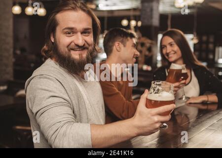 Bearded happy man smiling to the camera joyfully with a glass of beer in his hand, his friends chatting on the background. Cheerful man enjoying beer Stock Photo