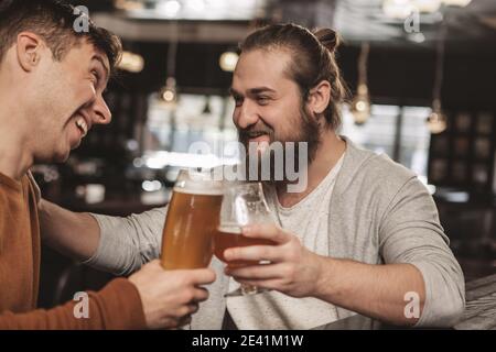 Two happy male friends laughing and talking over a glass of beer at the local pub. Young men relaxing, drinking delicious craft beer together at the b Stock Photo