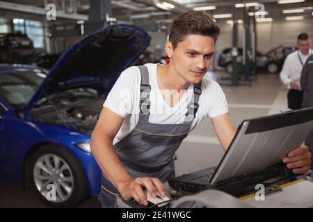 Handsome young male mechanic using computer, repairing automobile at the garage. Cars, transportation maintenance concept Stock Photo
