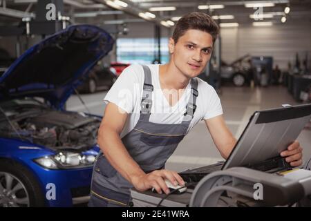 Young handsome car mechanic smiling to the camera, using laptop for computer diagnostics of a broken vehicle. Cheerful car technician working at the g Stock Photo