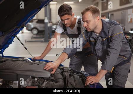 Mature male car mechanic helping his younger colleague repairing broken automobile. Two car service technicians looking under the hood of an auto Stock Photo