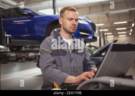 Mature male car mechanic looking concentrated, doing computer diagnostics on a broken automobile at his workshop. Cars, transportation concept Stock Photo